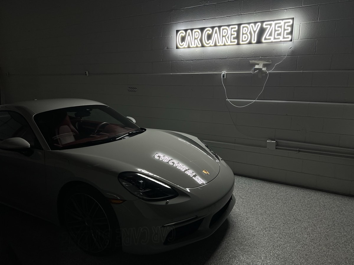 White Porsche 718 Cayman in the darkened shop, the Car Care by Zee neon sign reflected across its hood.